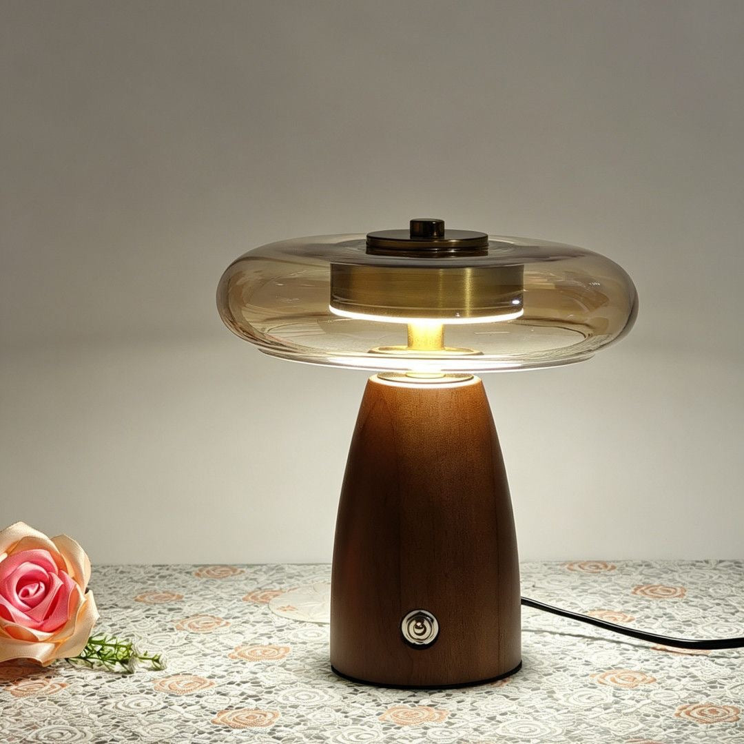 A mushroom-shaped table lamp with a dark brown wooden base and a clear glass cap, glowing with warm light. A pink rose is placed on the patterned tablecloth to the left, and a power cord extends to the right against a neutral gray background.