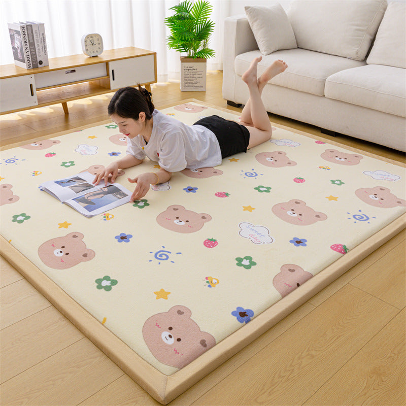 A woman lies on a large beige play mat with a light wood border in a bright living room. The mat is printed with cartoon teddy bears, flowers, stars, and small colorful motifs.