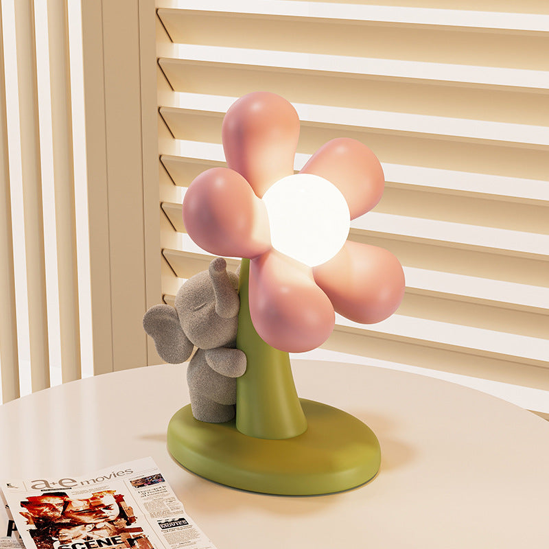 A cute flower-shaped table lamp with pink petals, a glowing white center, and a small gray elephant holding the stem, set on a round table in front of beige window blinds.