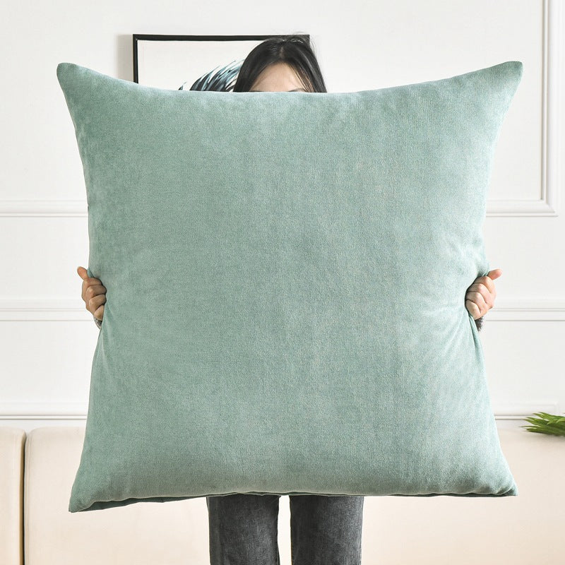 A person holds a large square throw pillow in a light sage green velvet fabric, standing in front of a white paneled wall.