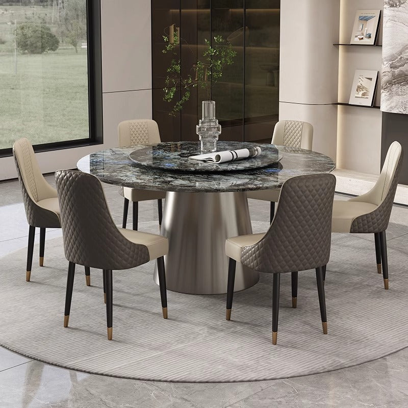 A modern round dining set featuring a dark marble-patterned table with a brushed metal pedestal base and six upholstered dining chairs in taupe and cream, arranged on a round gray rug in a contemporary dining room.