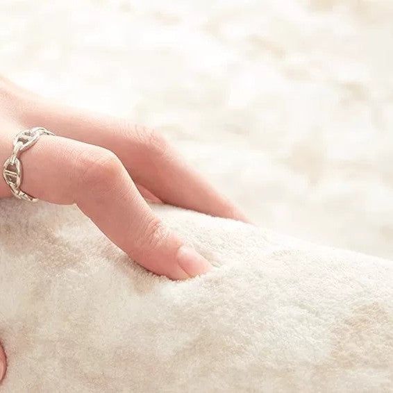 A close-up of a hand wearing a silver chain-link ring, gently resting on a soft, textured cream-colored plush surface.