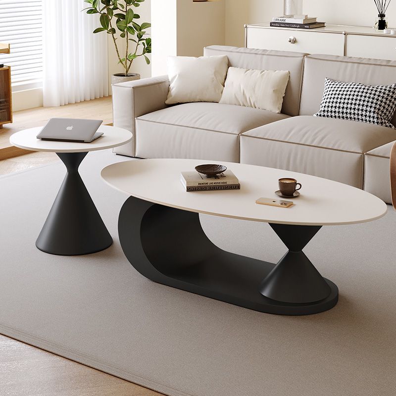Modern living room furniture set featuring an oval white coffee table with a sculptural black base, a matching round side table, and a light beige sofa in a bright, contemporary living room.