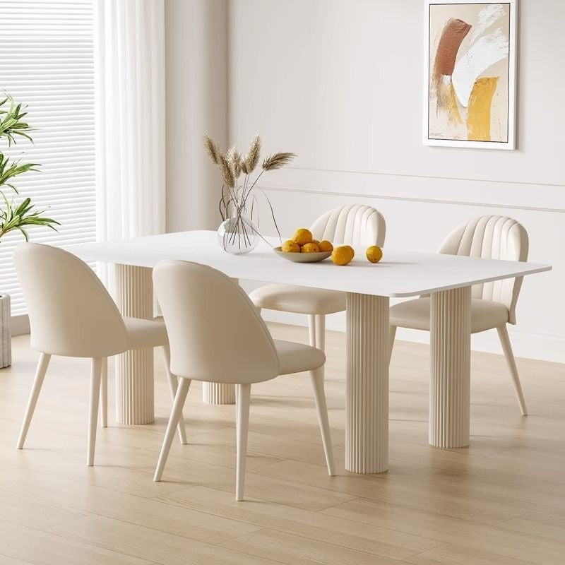 A modern dining set featuring a white rectangular table with fluted pedestal legs and four matching cream upholstered chairs, styled in a bright, minimalist dining room.