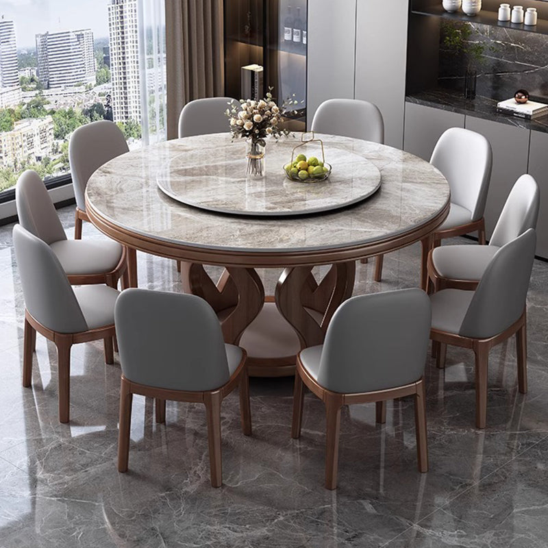A round dining table with a light marble-patterned top and carved wooden base, surrounded by eight gray upholstered chairs with wooden frames, set in a modern dining room with a large window view.
