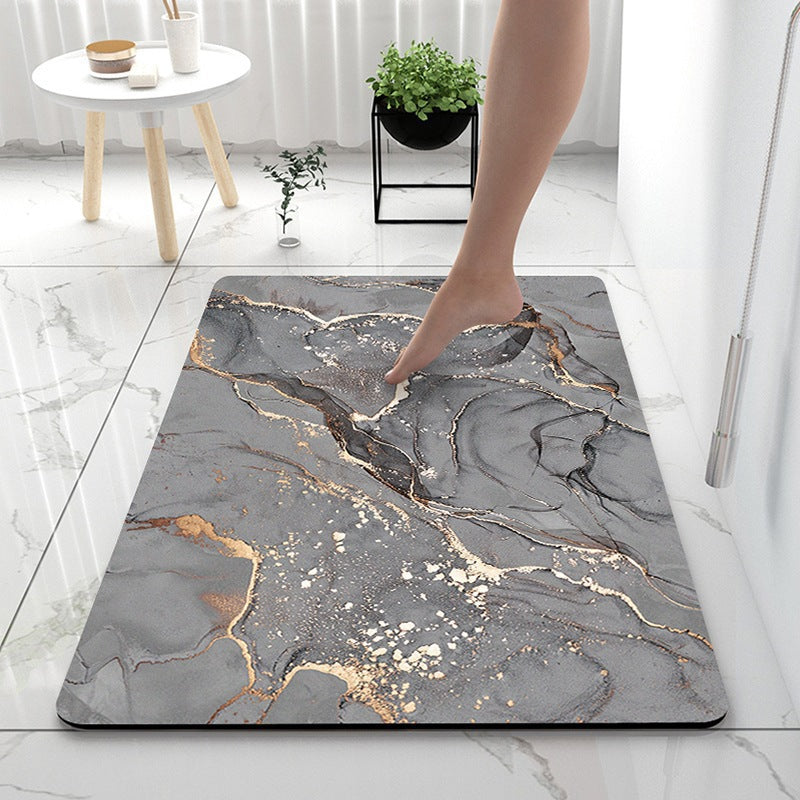 A rectangular bathroom mat with a gray marble pattern and metallic gold veining, shown on a white marble floor with a person's foot stepping onto it.