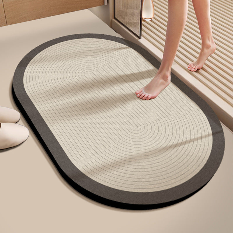 An oval entryway mat with a dark gray border and a beige center featuring concentric circular grooves, shown on a beige floor with a person's feet stepping onto it.