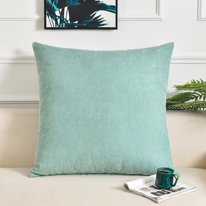 A square mint green throw pillow with a textured velvet-like fabric, styled on a beige sofa with a green mug, a magazine, and a framed print in the background.