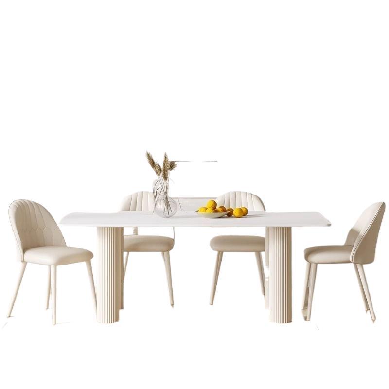 A modern dining set featuring a rectangular white table with fluted pedestal legs and four upholstered cream chairs, styled with a glass vase and lemons on a solid white background.
